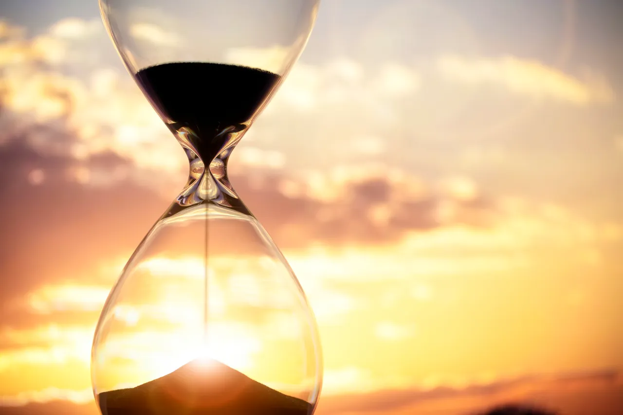 alt=An hourglass with sand flowing through it against a beautiful sunset backdrop.