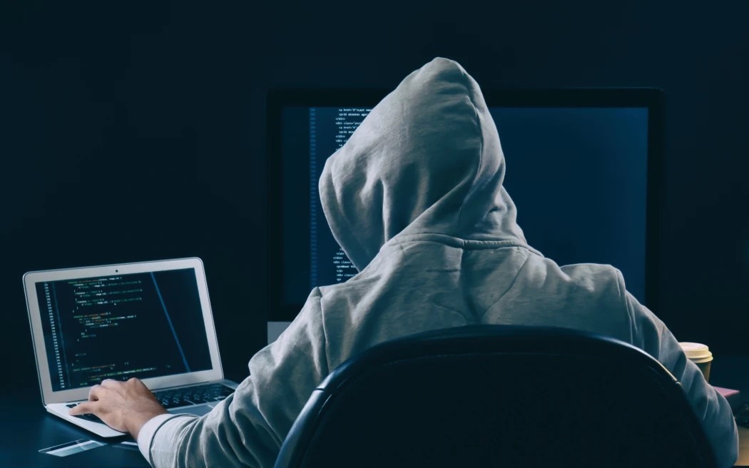 alt=A person in a hoodie sitting at a desk with a laptop, possibly involved in a data breach.