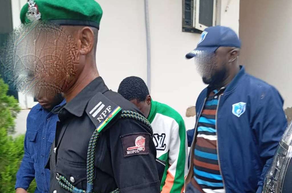 alt=A man in a green hat and shirt is led away by police officers, indicating a law enforcement action. Arrests in international operation targeting cybercriminals in West Africa
