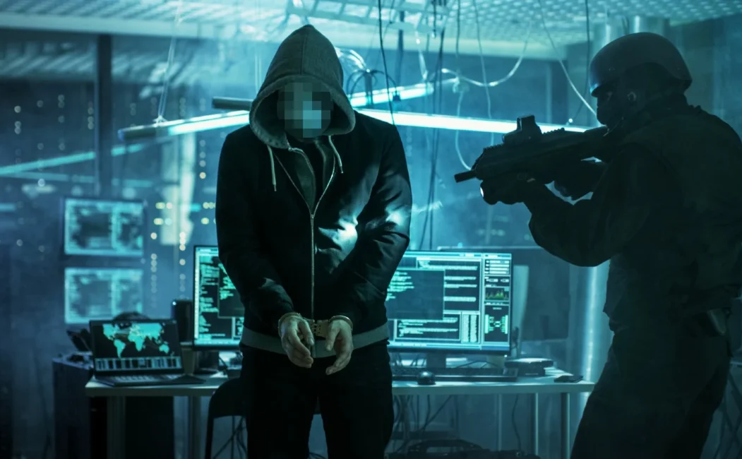 alt=A hooded figure stands with hands bound, while a tactical officer aims a weapon at them in a dimly lit, tech-filled room.