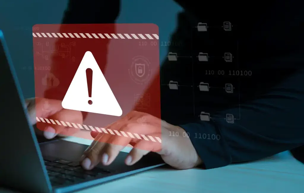 alt=A person typing on a laptop with a red warning symbol overlay, indicating a potential security threat or data breach.