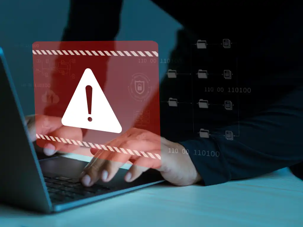 alt=A person typing on a laptop with a red warning symbol overlay, indicating a potential security threat or data breach.