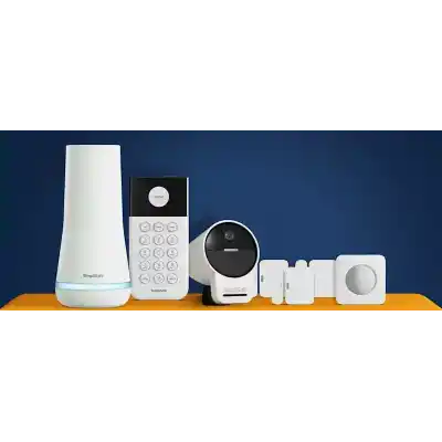 alt=A collection of SimpliSafe home security devices displayed on an orange surface against a blue background, including a hub, keypad, camera, and sensors.