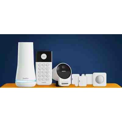 alt=A collection of SimpliSafe home security devices displayed on an orange surface against a blue background, including a hub, keypad, camera, and sensors.