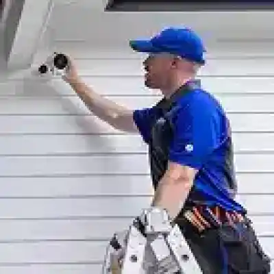 alt=A technician installs a security camera on a white wall, using a ladder and tool belt, wearing a blue uniform and cap.