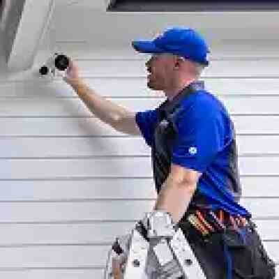 alt=A technician installs a security camera on a white wall, using a ladder and tool belt, wearing a blue uniform and cap.