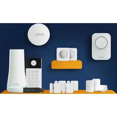 alt=A collection of home security devices on a vibrant orange tabletop, featuring sensors, a keypad, a hub, and a smoke detector against a dark blue wall.