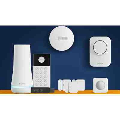 alt=A collection of SimpliSafe security devices on an orange surface, including a hub, keypad, sensors, and smoke detector against a blue background.