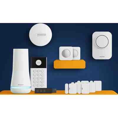 alt=A collection of home security devices, including a base station, keypad, sensors, and a smoke detector, on a vibrant blue and orange background.