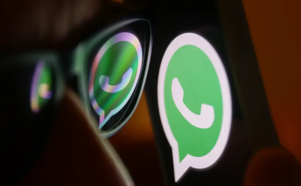 alt=A close-up of a person’s face wearing glasses, reflecting the green WhatsApp logo displayed on a smartphone screen.