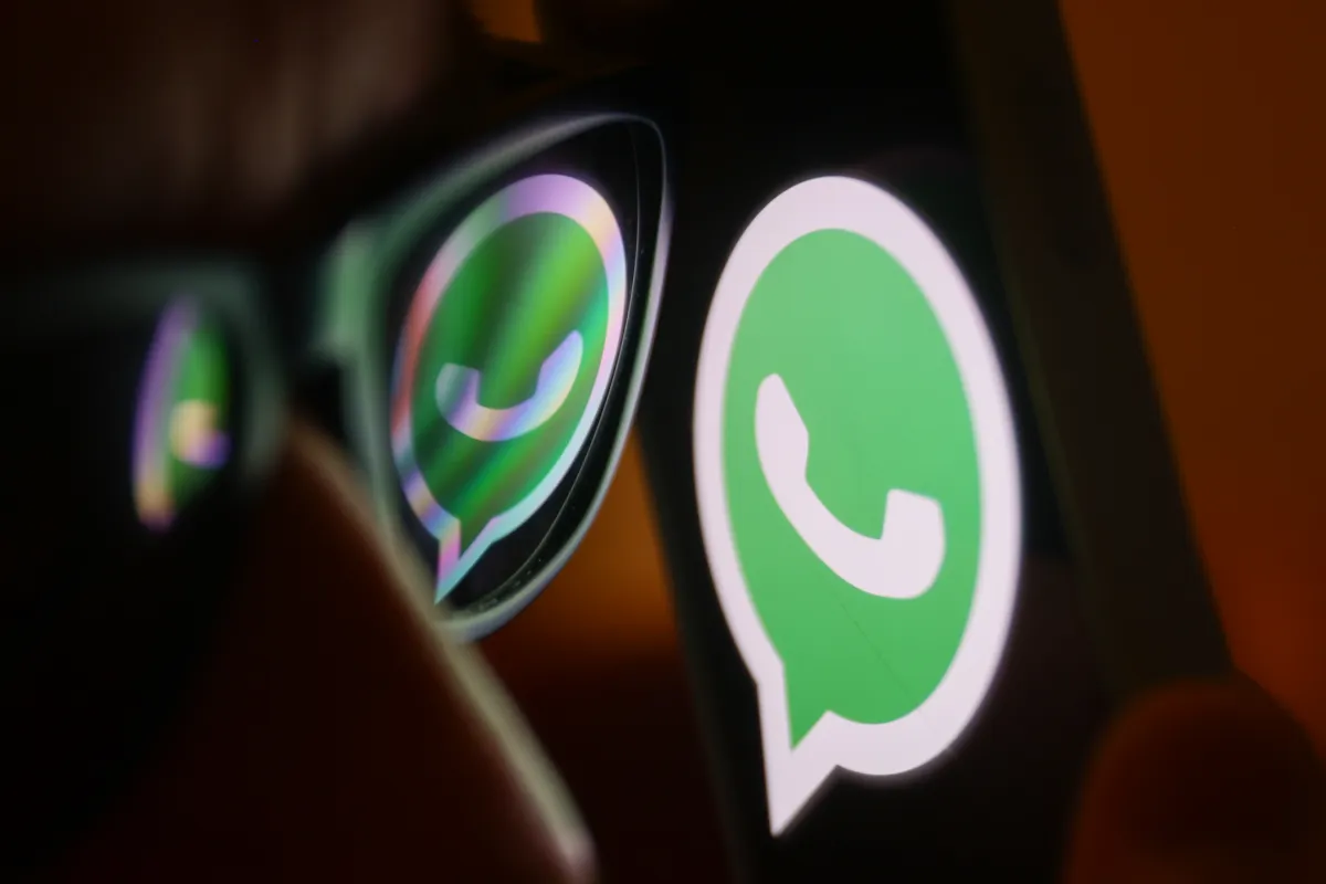 alt=A close-up of a person’s face wearing glasses, reflecting the green WhatsApp logo displayed on a smartphone screen.