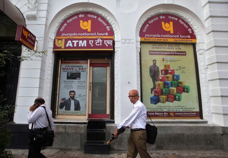 alt=Entrance of Punjab National Bank with ATM signage and two men walking past the building on a city street.
