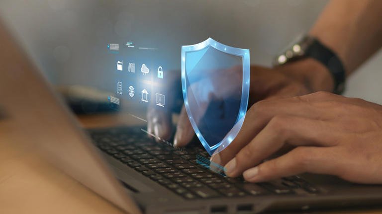 alt=Hands typing on laptop keyboard with a digital shield and security icons holographically displayed, symbolizing cybersecurity protection.